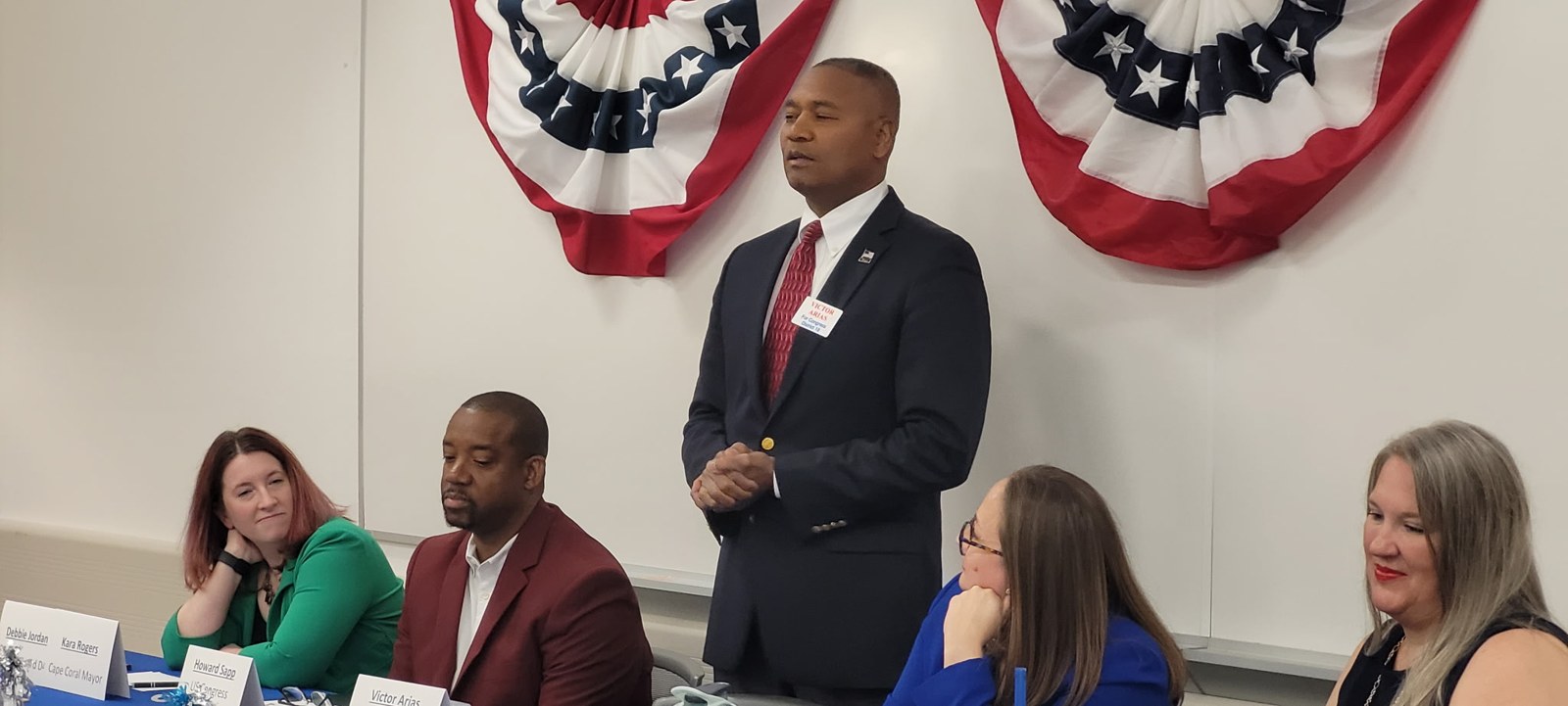 Victor Arias speaking at a FL-19 candidate forum panel with American flag bunting in the background