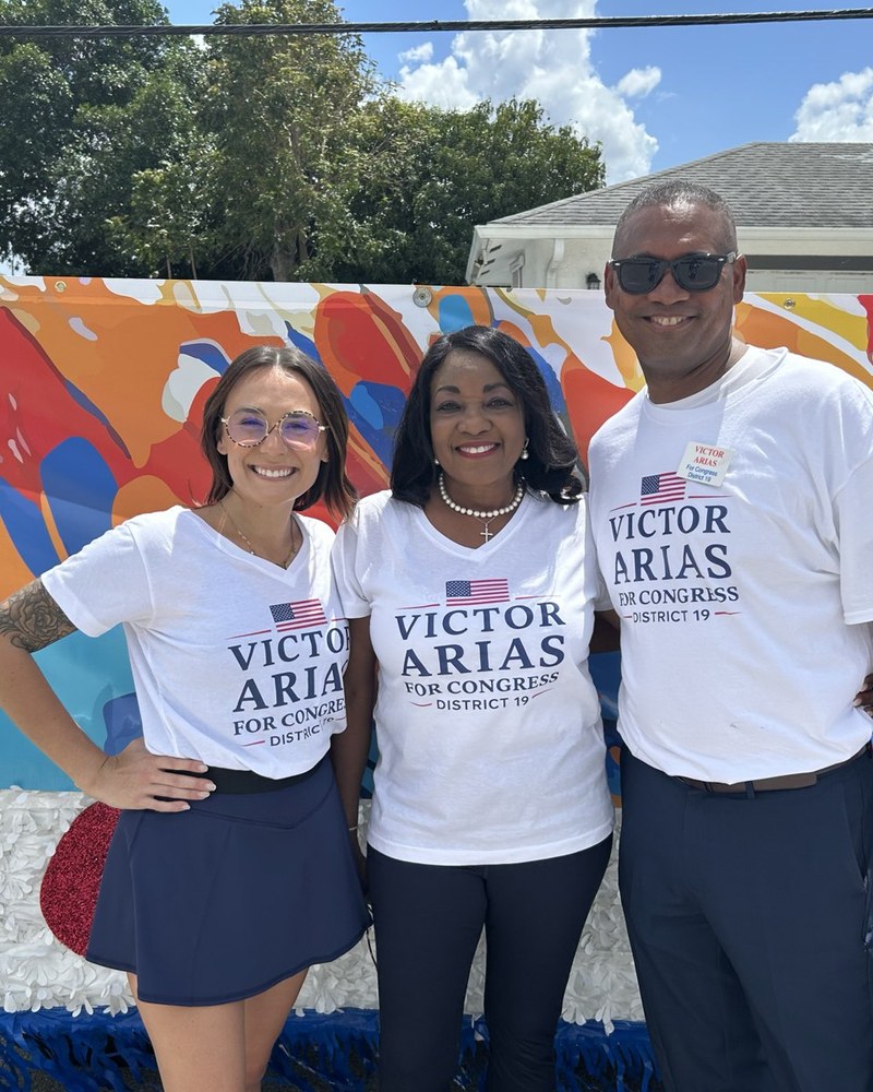 Victor Arias and two campaign volunteers smiling in matching Victor Arias for Congress t-shirts in front of a colorful mural at the Dunbar Easter Parade